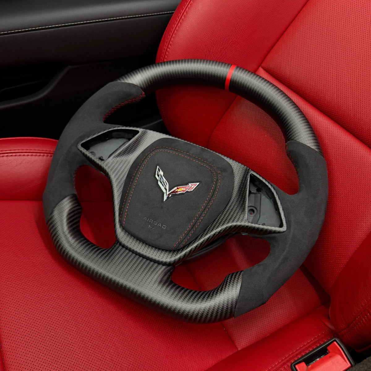 2014–2019 Chevrolet Corvette C7 woven carbon fiber flat-bottom steering wheel with black Alcantara grips, red stitching, red center stripe, and matching Alcantara airbag cover with crossed-flags emblem, resting on a red leather seat.